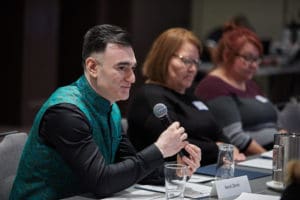 Photo of Harsh Zaran sitting at a table with a microphone surrounded by others who are listening